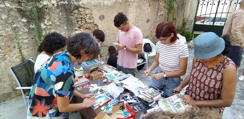 Image shows a courtyard with six people stood around a table full of tools and materials. The people are cutting out images and painting pieces of cardboard.