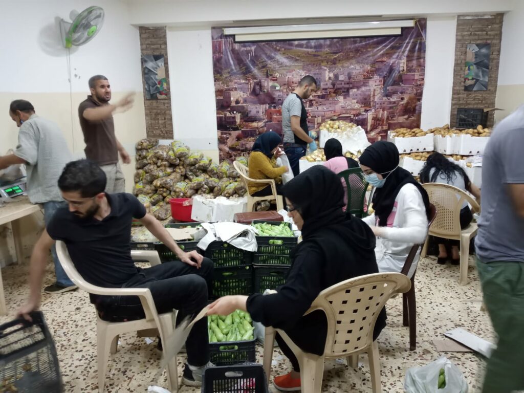 Residents of Bourj Albarajenah refugee camp are organising a food-sharing activity. There a people sat on chairs surrounding black plastic crates full of vegetables. People are preparing food ingredients, and are engaging in the process of food preparation in a collective manner. 
