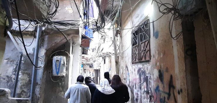 a street view from one of the passageways of Bourj albarajenah refugee camp. The camp is characterized by maze-like hallways which people often use as a public and social space. Looking towards the sky, you can see networks of electrical cables, but on the ground, you will see networks of people.