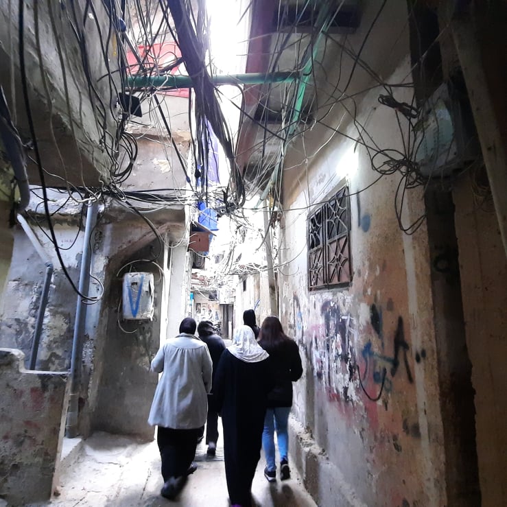 A street view from one of the passageways of Bourj albarajenah refugee camp. Towards the sky there are a mass of entangled electrical cables. There is a wall on one side of the passageway covered in graffiti. In the background there are several walking through the passageway.