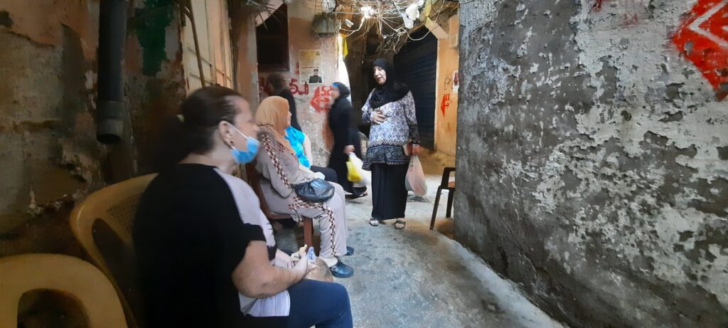 The image shows several women sat on plastic chairs in a passageway in the refugee camp. The pained concrete walls to the sides of the passageway appear old and worn. One woman is stood up talking to one sat down. Above, there are several entangled electrical cables. 