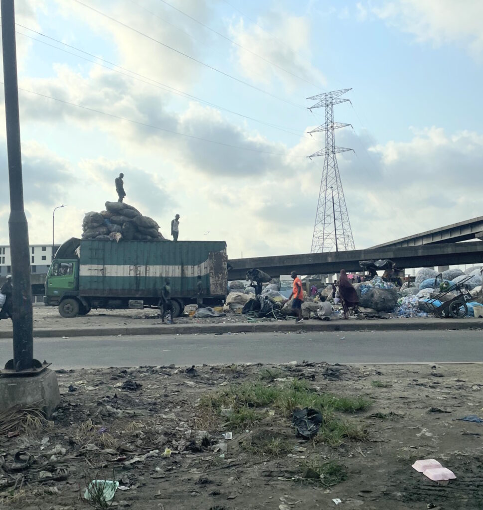 A sanitation and waste management hub in Lagos, where we see a large truck with stacks of refuse and two men standing on top of the truck, looking over piles of refuse. In the foreground, there is a road and a patch of ground with litter.