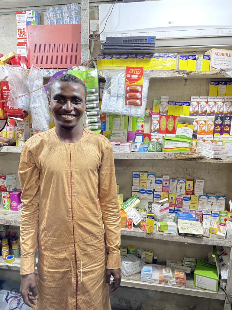 A young man wearing a long gold colored shirt is standing in front of shelves stocked with medications, with many packets of different-coloured packaging. He is smiling and looking at the camera.