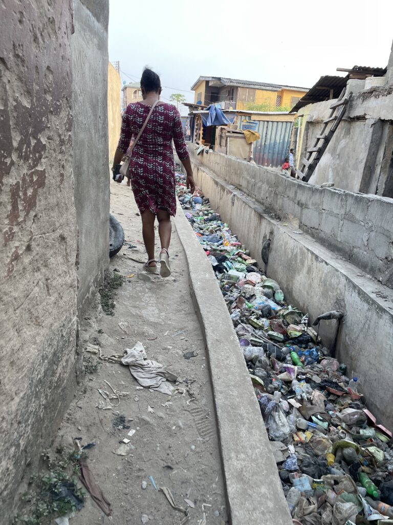 The back of a female health worker walking past a trench filled with refuse, next to small homes made of concrete and corrugated iron.
